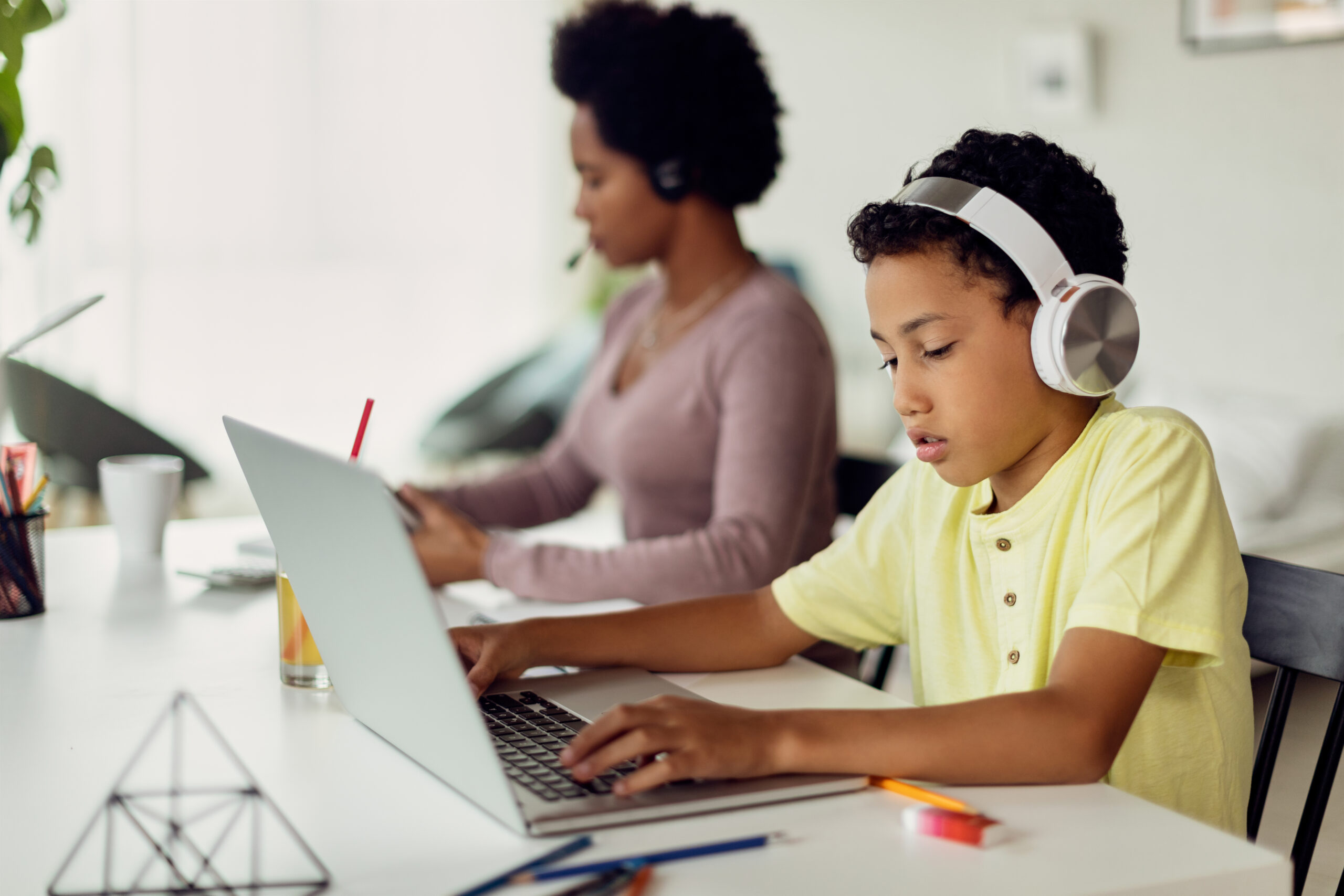 Small black boy with headphones surfing the net on a computer while his mother is working next to him at home.